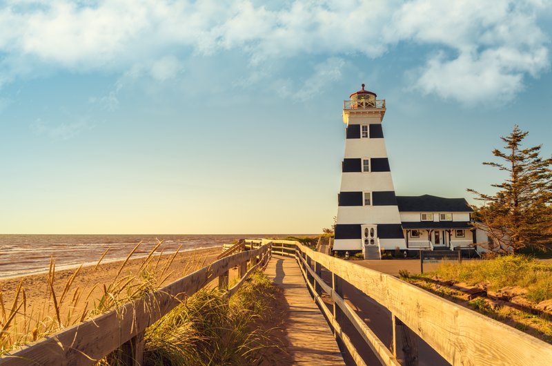 Striped West Point Lighthouse on Prince Edward Island at sunset with a wooden boardwalk leading to the beach