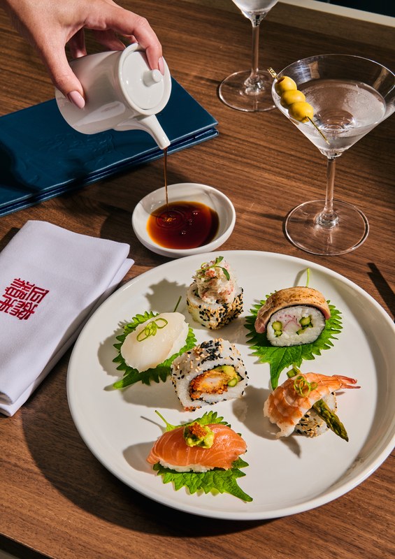 Morimoto Sushi Mori chef-selected sushi assortment on a white plate with soy sauce being poured, Morimoto by Sea napkin and a cocktail
