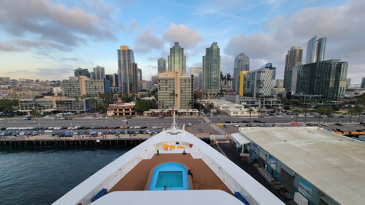 Cruise ship bow docked at the San Diego cruise terminal with the downtown skyline and Embarcadero behind it